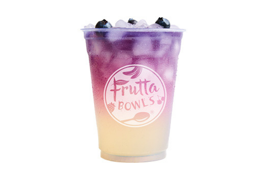 A Butterfly Refresher from Frutta Bowls, served in a clear cup with a gradient from pale yellow at the bottom to vibrant purple at the top, filled with ice cubes and topped with whole blueberries.