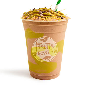 A Dubai Chocolate Smoothie from Frutta Bowls, served in a clear cup with light green swirl accents, topped with a drizzle of green nut butter, shredded toppings, and cacao nibs, with a green-striped straw inserted.
