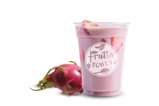 A Strawberry Dragonfruit Refresher in a clear Frutta Bowls cup, featuring a light pink drink with ice cubes and sliced strawberries, placed beside a whole vibrant pink dragon fruit with green-tipped scales.