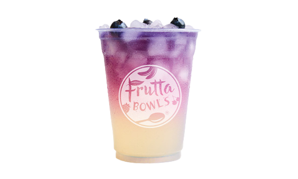 A cold, layered fruit drink in a plastic cup with the Frutta Bowls logo, featuring a gradient from pale yellow at the bottom to rich purple at the top, filled with ice cubes and garnished with whole blueberries.
